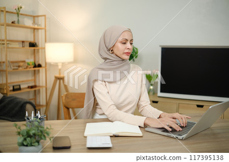 A woman wearing a scarf sits at a desk with a laptop and a notebook 117395138