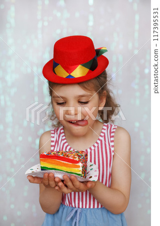 Portrait of happy surprised child girl of 6 years n festive red hat looks on piece of cake in rainbow colors on white plate in hands against festive backdrop Portrait of happy surprised child girl of 6 years n festive red hat looks on piece of cake in rainbow colors on white plate in hands against festive backdrop 117395531