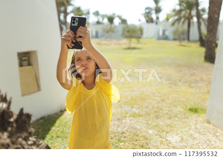 Girl 7 years old in yellow dress takes photos on phone on lawn with palm trees in summer day 117395532