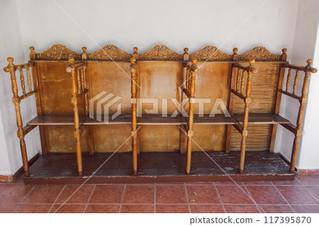 A set of empty, vintage wooden church chairs with intricate carvings arranged outdoors against a white wall and floor with reddish-brown tiles. 117395870