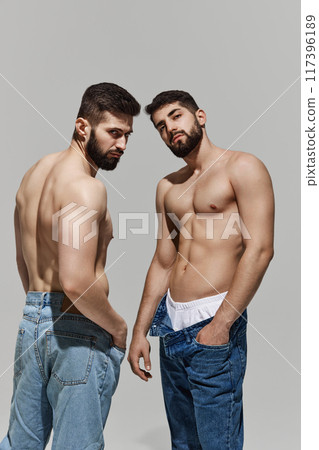 Two shirtless men stand confidently in jeans, they intense looks at camera, exuding strength and attitude against warm grey background. Two shirtless men stand confidently in jeans, they intense looks at camera, exuding strength and attitude against warm grey background. 117396189