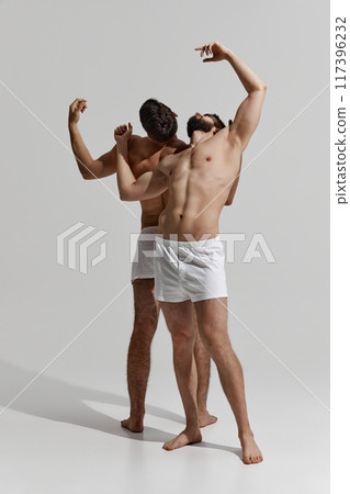 Two men in white underwear strike expressive poses, their bodies intertwined in moment of fluid motion against warm grey background. Two men in white underwear strike expressive poses, their bodies intertwined in moment of fluid motion against warm grey background. 117396232