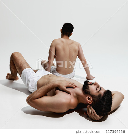 Two men in white shorts relax, one lying down with hand on his chest, other sitting upright, both reflecting moment of calm and rest. Two men in white shorts relax, one lying down with hand on his chest, other sitting upright, both reflecting moment of calm and rest. 117396236