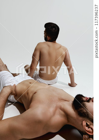 Two men in white shorts relax, one lying down with hand on his chest, other sitting upright, both reflecting moment of calm and rest. Two men in white shorts relax, one lying down with hand on his chest, other sitting upright, both reflecting moment of calm and rest. 117396237