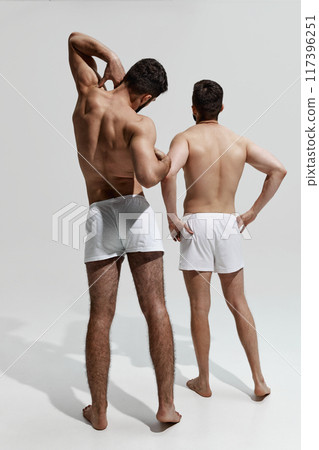 Rear view photo of two young men posing in white underwear against warm grey background. Healthy back. Rear view photo of two young men posing in white underwear against warm grey background. Healthy back. 117396251