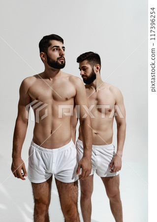 Two brutal male models with beards, with muscular build bodies posing shirtless, in pants against warm grey background. 117396254