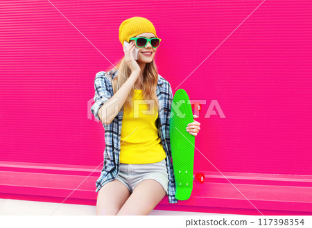 Stylish smiling young woman calling on phone with skateboard in bright yellow hat on pink background 117398354
