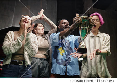 Group of diverse young people celebrating victory while holding a trophy in cozy indoor setting with joyful expressions, raising arms in excitement 117398465