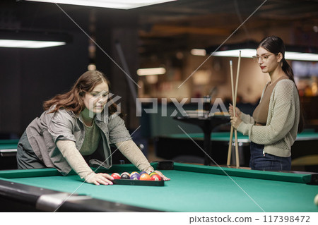 Two women getting ready for billiards game in a modern recreation center with dim lighting. One woman setting up balls, the other holding cue stick Two women getting ready for billiards game in a modern recreation center with dim lighting. One woman setting up balls, the other holding cue stick 117398472