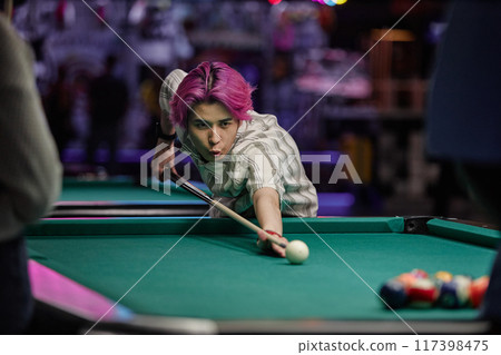 Young person with pink hair intensely focusing on taking shot while playing pool in brightly lit recreational venue featuring vibrant colors and various entertainment facilities 117398475