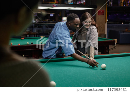 African American man leaning over billiard table with Caucasian woman, aiming for a shot while smiling and engaging in the game in a recreational setting African American man leaning over billiard table with Caucasian woman, aiming for a shot while smiling and engaging in the game in a recreational setting 117398481