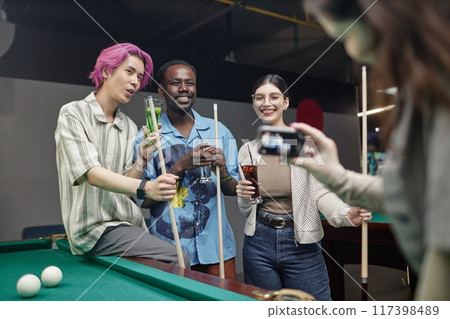 Group of young friends smiling and enjoying playing pool while holding drinks in hand, person photographing them. Happy and casual atmosphere with green pool table in foreground Group of young friends smiling and enjoying playing pool while holding drinks in hand, person photographing them. Happy and casual atmosphere with green pool table in foreground 117398489