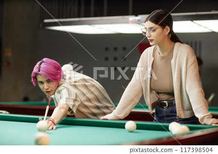 Young man with pink hair aiming billiard cue while woman with glasses watching attentively in billiard hall having some focus 117398504