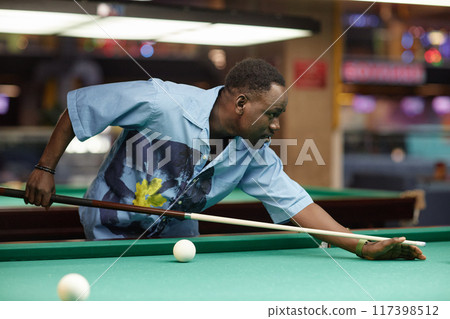 Man aiming pool cue while focusing on billiard table in dimly lit recreational space. Casual attire and colorful background contribute to lively atmosphere Man aiming pool cue while focusing on billiard table in dimly lit recreational space. Casual attire and colorful background contribute to lively atmosphere 117398512