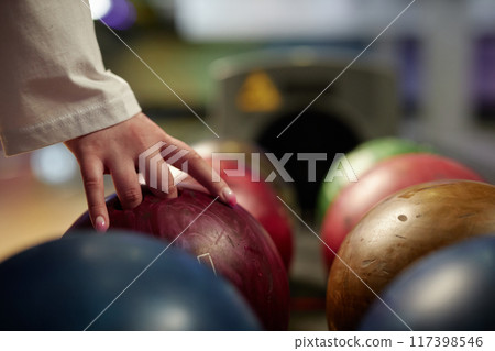 Hand reaching for vibrant bowling ball in colorful, well-lit bowling alley with various balls and blurred background creating dynamic atmosphere Hand reaching for vibrant bowling ball in colorful, well-lit bowling alley with various balls and blurred background creating dynamic atmosphere 117398546