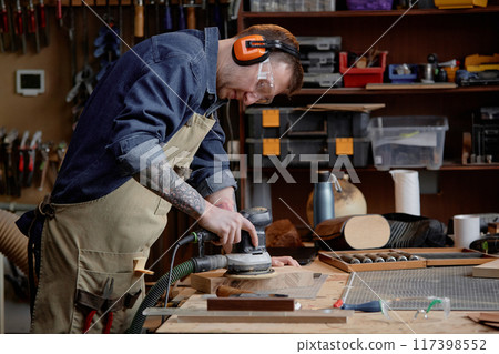 Artisan focusing on sanding wooden piece using electric sander in well-equipped woodworking workshop with various tools and materials arranged on shelves 117398552