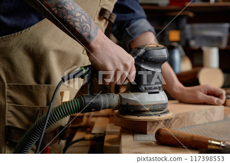 Detailed image showing craftsman using electric sander on wooden piece in workshop. Forearm with tattoo visible, emphasizing intricate work on smoothening wood surface Detailed image showing craftsman using electric sander on wooden piece in workshop. Forearm with tattoo visible, emphasizing intricate work on smoothening wood surface 117398553