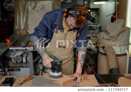Person seen concentrating on sanding wood piece with sander machine in workshop filled with various tools and gadgets surrounding. Wearing protective gear and focused on task Person seen concentrating on sanding wood piece with sander machine in workshop filled with various tools and gadgets surrounding. Wearing protective gear and focused on task 117398557