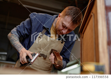 Woodworker concentrating on sanding wooden furniture piece, wearing apron and surrounded by workshop tools and equipment. Tattoed arms visible while engaged in meticulous craftsmanship Woodworker concentrating on sanding wooden furniture piece, wearing apron and surrounded by workshop tools and equipment. Tattoed arms visible while engaged in meticulous craftsmanship 117398558