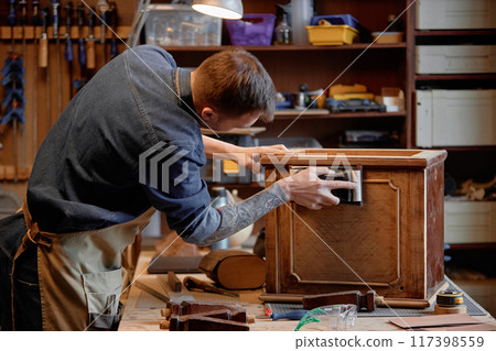 Man crafting intricate antique furniture piece in woodworking workshop surrounded by tools and materials, meticulously sanding the wood surface 117398559