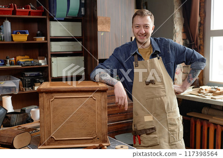 Smiling male craftsman wearing apron working on wooden cabinet in workshop surrounded by tools and materials creating woodwork project Smiling male craftsman wearing apron working on wooden cabinet in workshop surrounded by tools and materials creating woodwork project 117398564