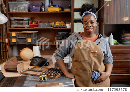 Young woman standing in front of workbench with various tools and supplies, holding a piece of cloth, and smiling confidently in a woodshop filled with crafting materials 117398578