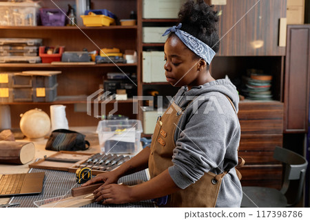 African American woman wearing apron and headband focused on her project in a craft workshop. Surrounding tools and materials suggest a hands-on creative process 117398786