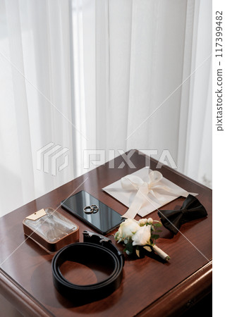 Groom's wedding essentials on table, featuring rings on phone, bow tie, boutonniere, perfume, belt Groom's wedding essentials on table, featuring rings on phone, bow tie, boutonniere, perfume, belt 117399482