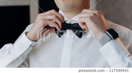 Groom adjusting his black bow tie, symbolizing elegance and style on his wedding day Groom adjusting his black bow tie, symbolizing elegance and style on his wedding day 117399491