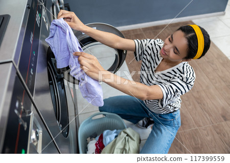 Woman in striped shirt doing routine laundry tasks at self-service laundromat 117399559