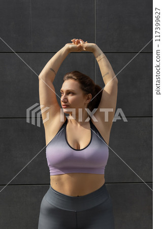 Plus size young woman in sports wear standing with her arms up and looking aside 117399627