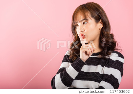 Portrait of a happy Asian woman making a quiet gesture by placing her finger on her lips. Studio shot isolated on pink, with space for your text. 117403602