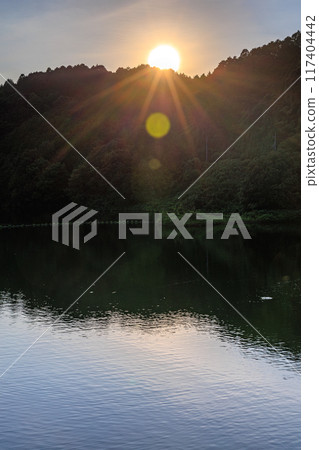 Summer at Nunome Dam in the eastern part of Nara City. Evening view towards the dam lake. The surface of the dam lake illuminated by the setting sun on the opposite bank. Summer at Nunome Dam in the eastern part of Nara City. Evening view towards the dam lake. The surface of the dam lake illuminated by the setting sun on the opposite bank. 117404442