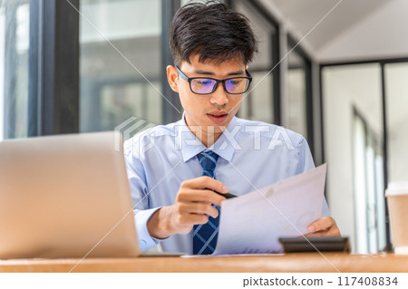 Young businessman working on his project in his office checking business result data from the printed sheets and his laptop computer Young businessman working on his project in his office checking business result data from the printed sheets and his laptop computer 117408834