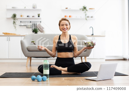 Smiling woman practicing yoga while eating healthy salad at home Smiling woman practicing yoga while eating healthy salad at home 117409008