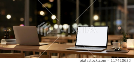 A laptop computer mockup on a wooden table in a contemporary coffee shop co-working space at night. 117410623