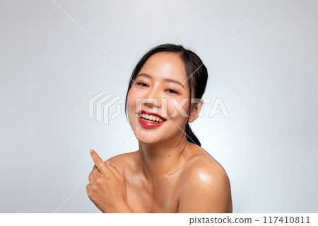 A pretty Asian woman smiles with her teeth visible while posing against a grey studio background. 117410811
