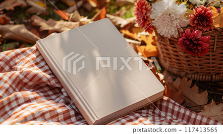 Picnic in an Autumn park. Beautiful scotch pattern blanket is laid on the ground with a book with blank white cover and a basket full of flowers beside. AI generated. 117413565