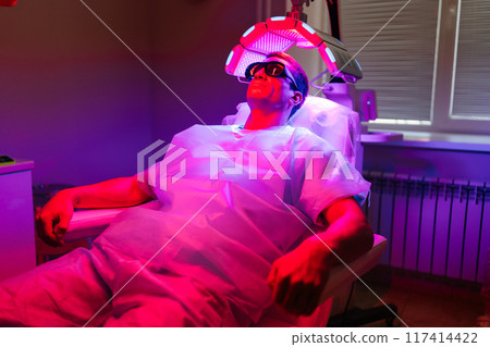 Relaxed handsome young man client with hairloss problem having LED light hair photodynamic therapy treatment after hair transplant at clinic. Male patient having non-invasive type of phototherapy Relaxed handsome young man client with hairloss problem having LED light hair photodynamic therapy treatment after hair transplant at clinic. Male patient having non-invasive type of phototherapy 117414422