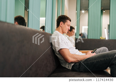 Relaxed male traveler in casual clothes relaxing in hotel lobby using smartphone watching online video or browsing internet. Male tourist resting with phone in lounge area at luxury resort. Relaxed male traveler in casual clothes relaxing in hotel lobby using smartphone watching online video or browsing internet. Male tourist resting with phone in lounge area at luxury resort. 117414430