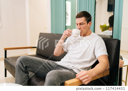 Successful male client sipping from cup of coffee while comfortably waiting in modern and welcoming lobby of medical clinic. Good looking male client enjoying cup of tea while relaxing in waiting room Successful male client sipping from cup of coffee while comfortably waiting in modern and welcoming lobby of medical clinic. Good looking male client enjoying cup of tea while relaxing in waiting room 117414431