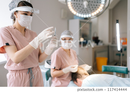 Portrait of gloved female nurse prepares filled syringe by tapping syringe and giving to doctor for injection. Anesthesiologist in mask removing air bubbles from syringe by tapping it. Portrait of gloved female nurse prepares filled syringe by tapping syringe and giving to doctor for injection. Anesthesiologist in mask removing air bubbles from syringe by tapping it. 117414444