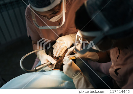 Close-up of two female doctors in lighting glasses extracting hair follicles from donor area of male patient for hair transplantation in clinic. Process of hair transplant with FUE Sapphire method. 117414445