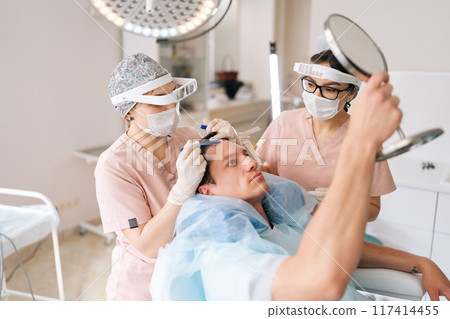 Medium shot of male patient with hair loss problem talking with female trichologist marking who with pencil new hairline on head before hair transplantation surgery. Concept of baldness treatment 117414455
