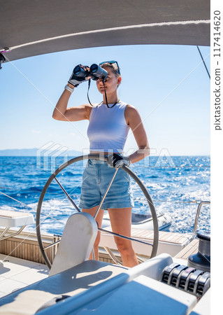 Young female captain on the yacht looking through binoculars during sailing boat control. Travel and active life. Yachting Young female captain on the yacht looking through binoculars during sailing boat control. Travel and active life. Yachting 117414620