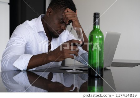 Stressed businessman sitting at kitchen counter table with laptop, wine glass and bottle. African man in white shirt rubbing forehead and looking troubled with hard working at home. 117414819