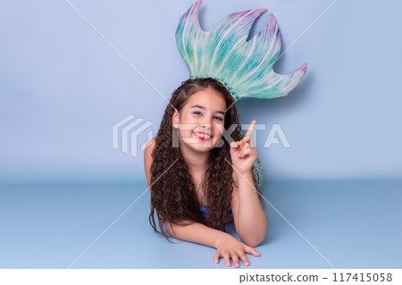 A happy child girl in a mermaid costume lie on a blue background A happy child girl in a mermaid costume lie on a blue background 117415058
