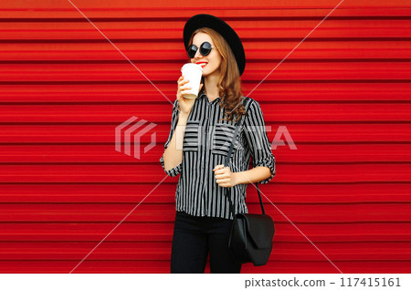 Stylish young woman with coffee cup in black shirt, round hat, handbag clutch on red wall background 117415161