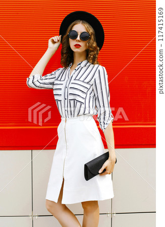 Beautiful young woman model wearing white striped shirt, black handbag clutch, round hat, skirt in the city 117415169