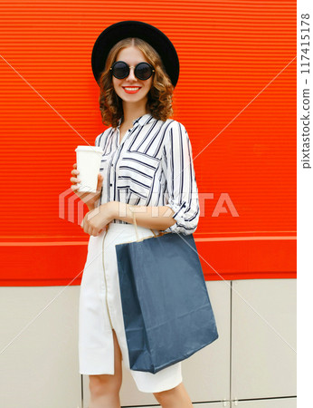 Beautiful young smiling woman with coffee cup and shopping bags in white shirt, black hat in the city Beautiful young smiling woman with coffee cup and shopping bags in white shirt, black hat in the city 117415178